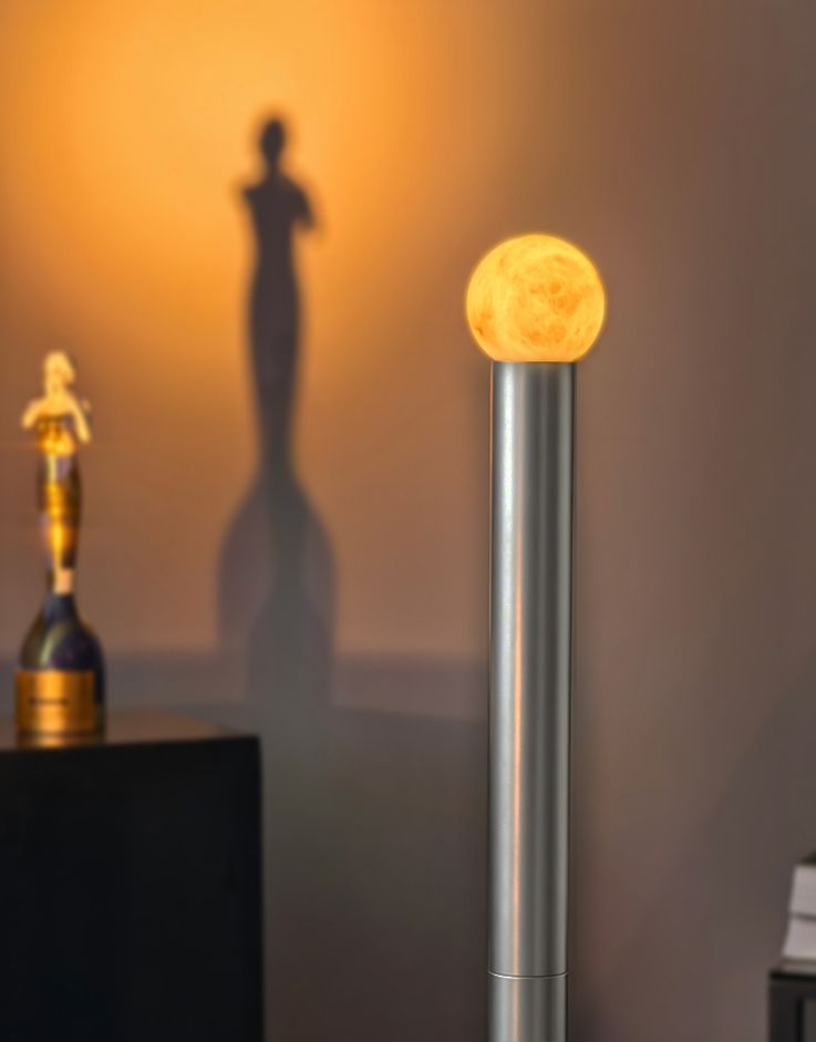 Moon-like glowing orb atop a silver lamp against a warm wall, casting shadows of statues in the background (decorative lamp).