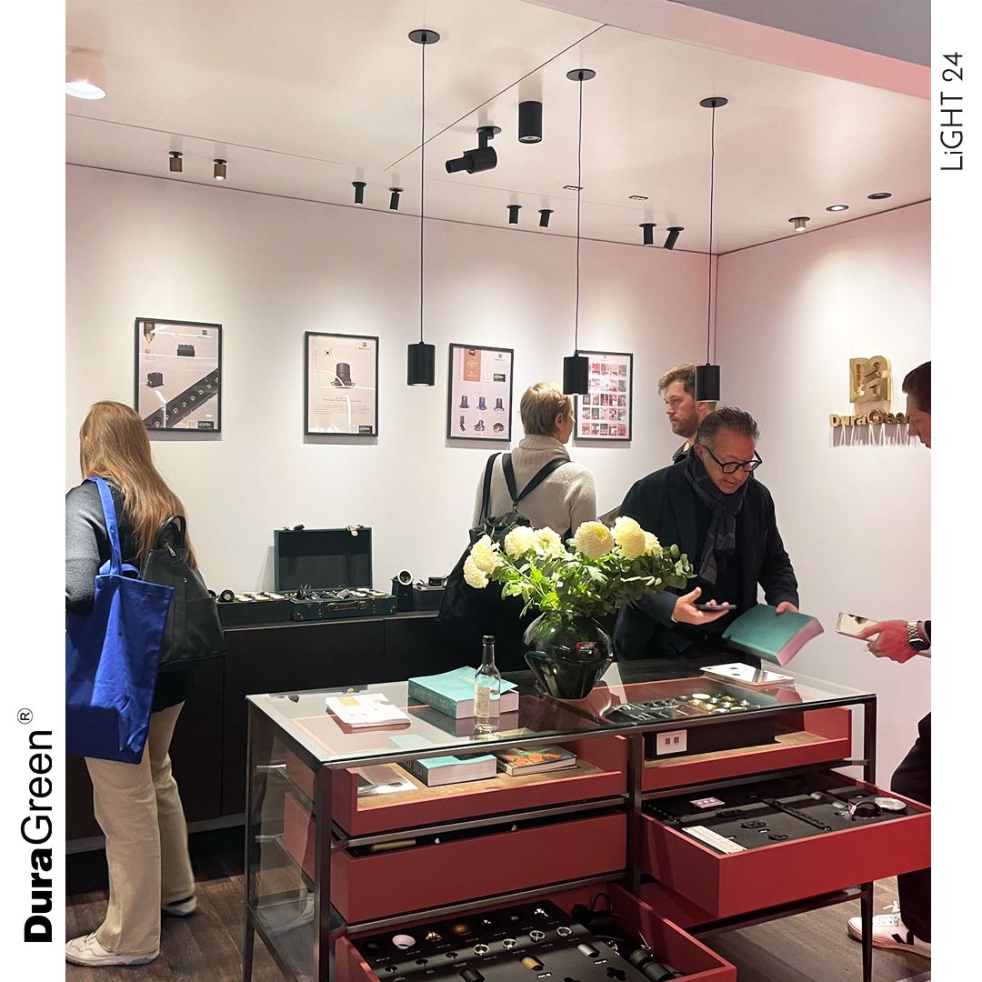 Shoppers browse a boutique with glass display tables, a vase of white flowers, and framed product posters on white walls.