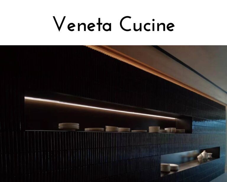 Logo reading 'Veneta Cucine' above a dark kitchen display with vertical tile backsplash and stacked plates on shelves behind glassed counters.