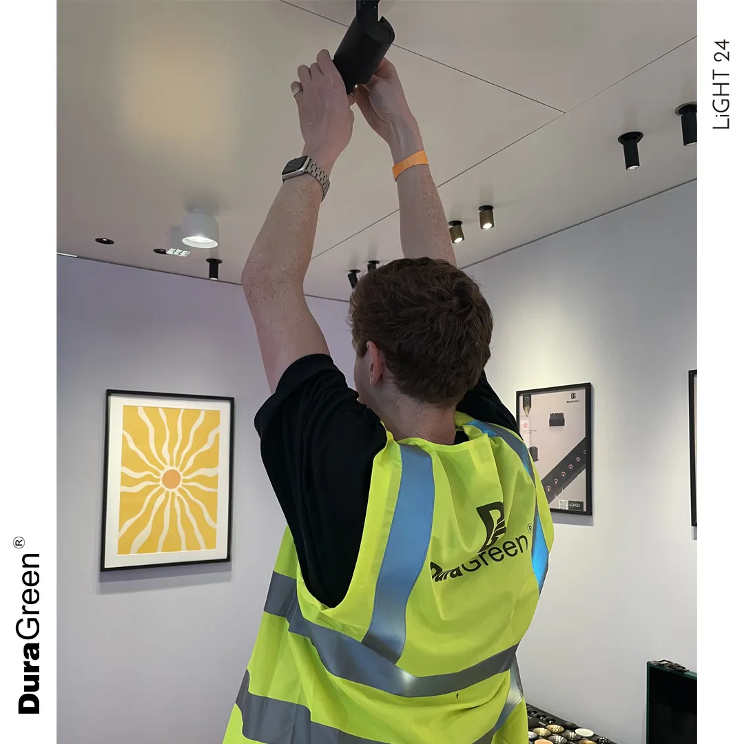 Worker in a neon safety vest installs or adjusts a ceiling light fixture in a gallery/space, arms raised overhead.