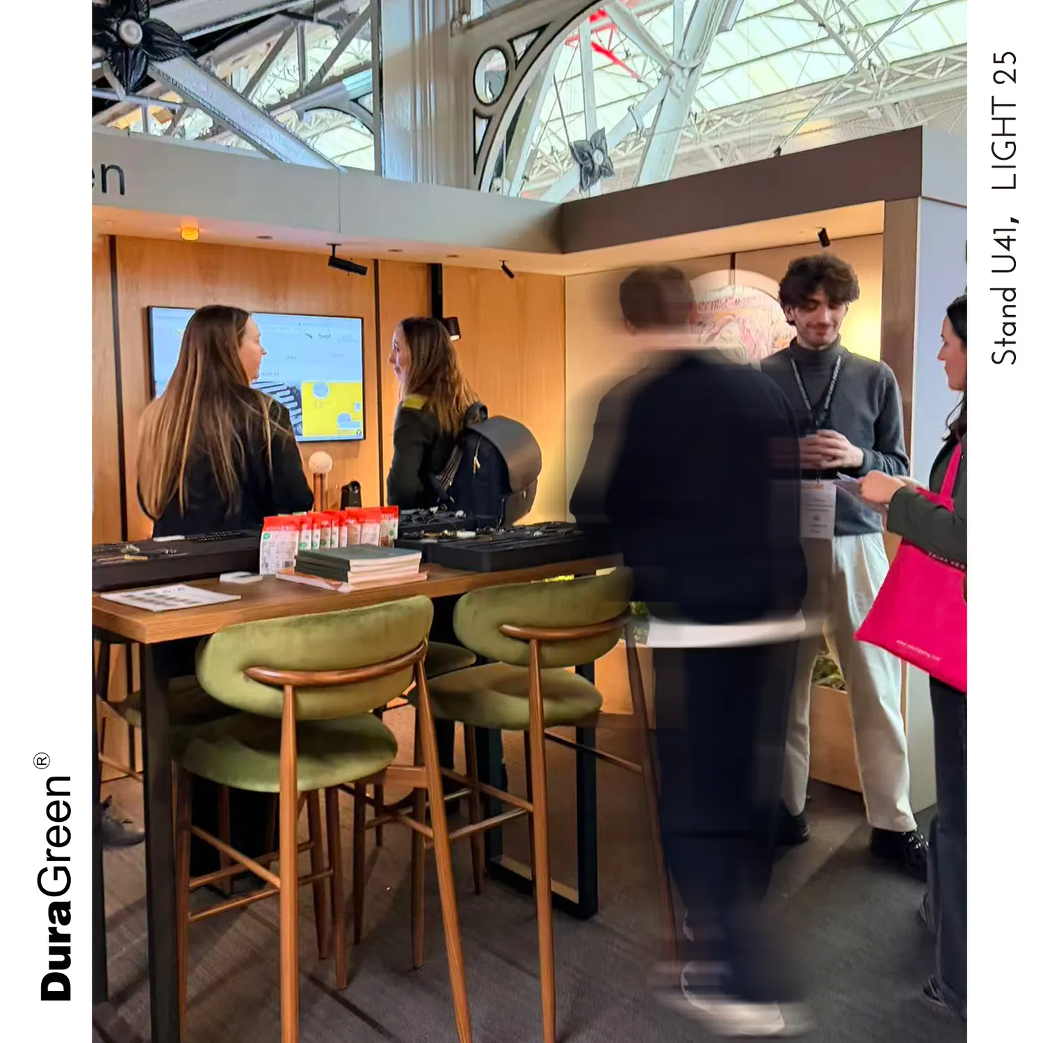 People chat at a trade show booth with a monitor, wooden backdrop, and a high table with green stools; brochures on the table.