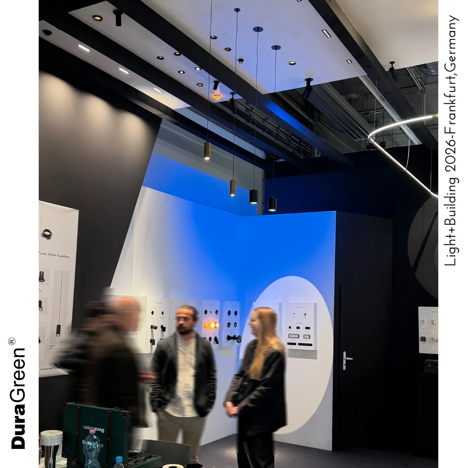 Exhibition booth with multiple pendant lights hanging from a ceiling, white and blue curved panels, and blurred visitors in front; vertical event branding along the sides.